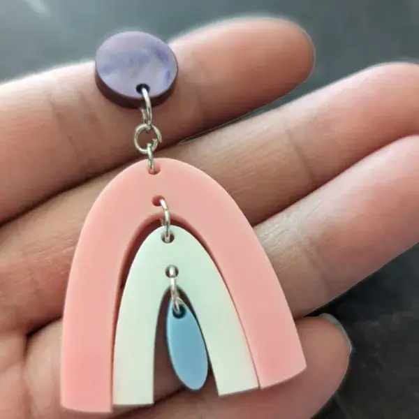 A pair of purple stud earrings dangling a pink rainbow, dangling a white rainbow, dangling a blue rainbow