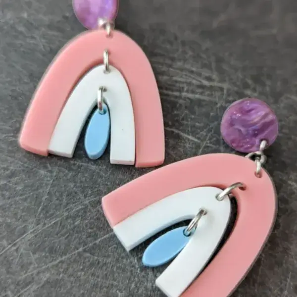 A pair of purple stud earrings dangling a pink rainbow, dangling a white rainbow, dangling a blue rainbow