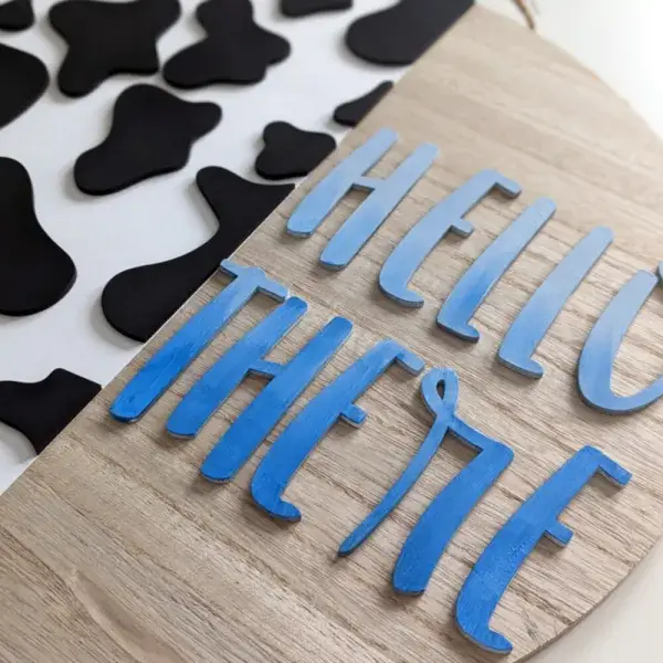 A round sign with jute hanger. The left hand side has cow print, the right says "Hello There"