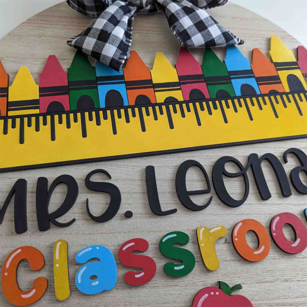 Detailed view of a round classroom sign for Mrs. Leonard with colorful crayon shapes, a ruler design, black script text, and a buffalo plaid bow.