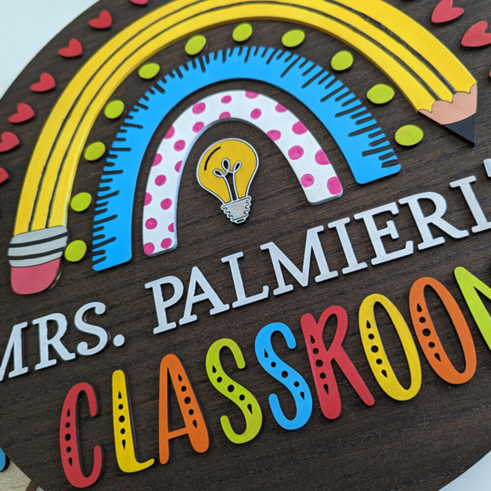 Close-up of a personalized classroom sign for Mrs. Palmieri with a rainbow made of pencil, ruler, and polka dot arches, featuring a light bulb at the center.