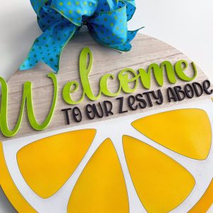 Close-up of a citrus-themed welcome sign with raised green 'Welcome' lettering, 'to our zesty abode' text, and a vibrant lemon wedge design.