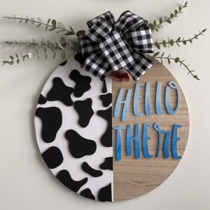 Rustic cow print welcome sign with blue 'HELLO THERE' text, accented with black-and-white buffalo check bow and eucalyptus greenery.