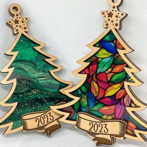 Two wooden Christmas tree ornaments side by side. The left tree has a green marbled acrylic background, and the right features a colorful stained-glass style leaf pattern. Both have engraved wooden stars at the top and banners with “2023” at the bottom.