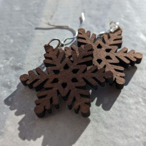 Pair of dark wood snowflake-shaped earrings with intricate cutout details and silver hooks, displayed on a light stone surface in natural sunlight.