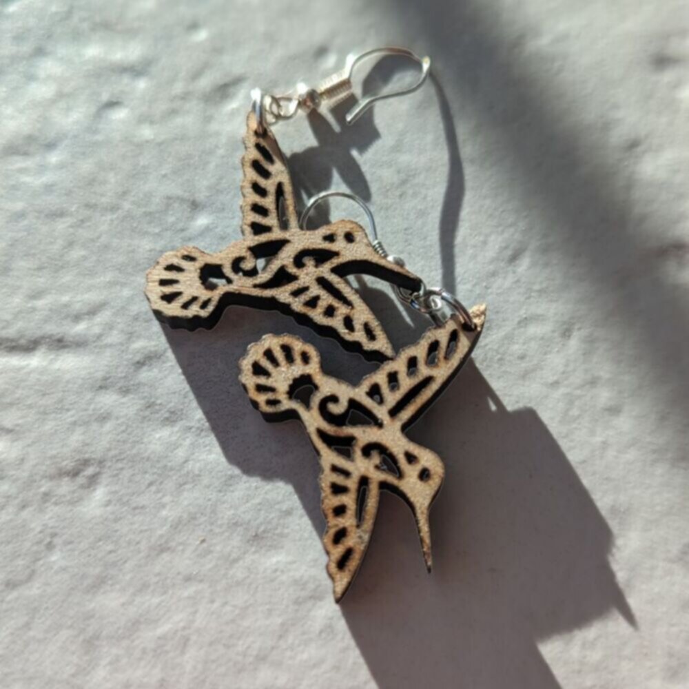 Wooden hummingbird earrings with intricate cutout design casting a shadow on a textured surface.