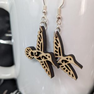 Pair of laser-cut wooden hummingbird earrings hanging on a white mug, shown in close-up.