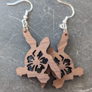 Pair of wooden sea turtle earrings with laser-cut hibiscus flowers, shown on a dark stone background.