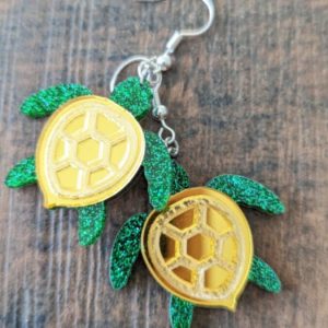 Close-up of turtle earrings with hexagonal yellow shells and sparkly green limbs on a wood texture.