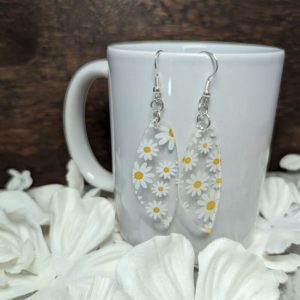 Daisy-themed acrylic earrings hanging from a white ceramic mug, surrounded by white flower petals on a wooden background.
