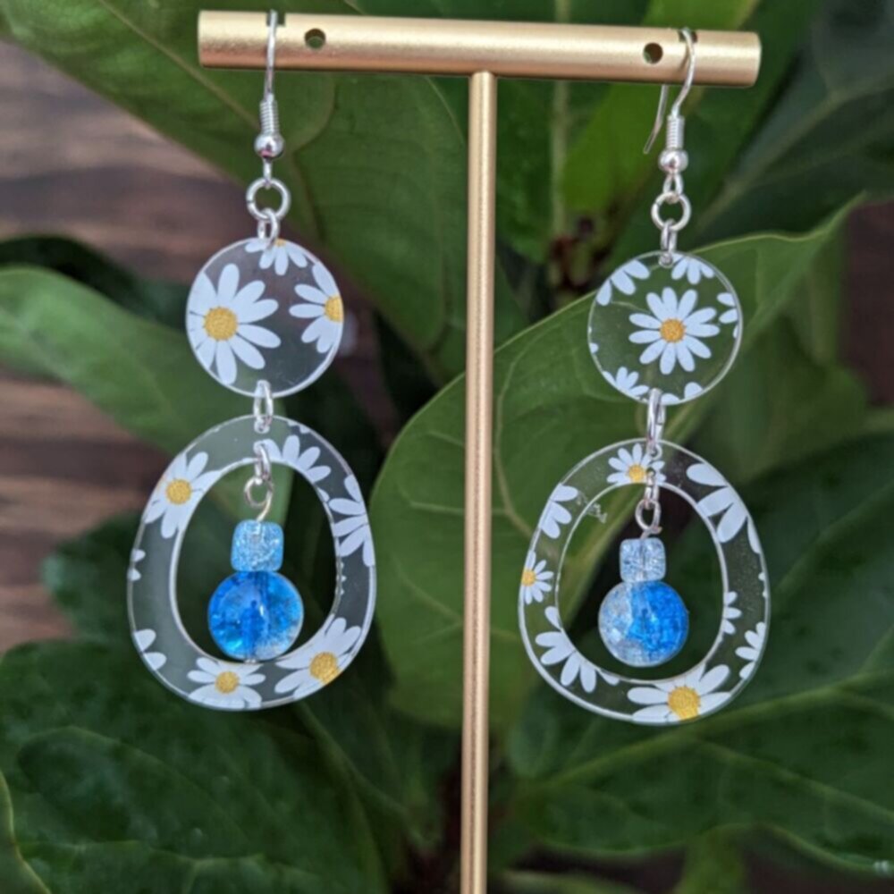 Dangle earrings with clear circular and oval daisy-print acrylic charms, accented by shimmering blue glass beads, displayed on a gold earring stand in front of green leaves.