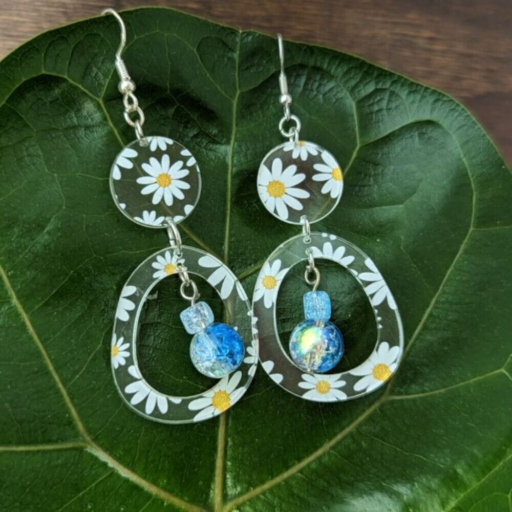 Pair of floral acrylic earrings with daisy patterns and dangling iridescent blue beads, laid flat on a large green leaf.