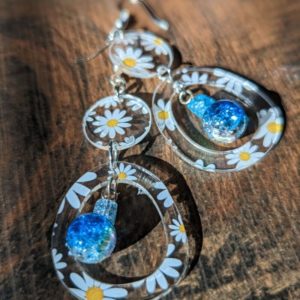 Close-up of daisy-themed acrylic earrings with round blue beads, resting on a wooden surface in bright natural light.