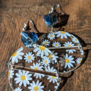 Earrings with bold daisy print and blue gemstone tops laid on a rustic wood table with dramatic sunlight and shadows.