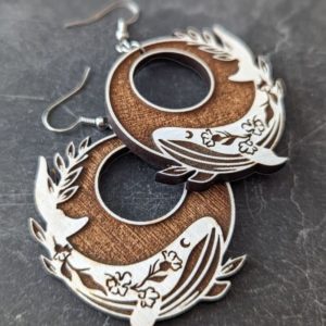 Handcrafted circular earrings featuring white dove and floral engraving displayed on a dark stone surface.