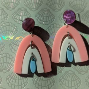 Pair of pastel rainbow earrings displayed on a textured background with embossed shell patterns, illuminated by sunlight, showing pink, white, and blue arcs with purple studs.