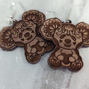 Close-up of two wooden bear-shaped earrings with detailed etching, featuring floral ears and heart-shaped accents, placed on a light marble surface.