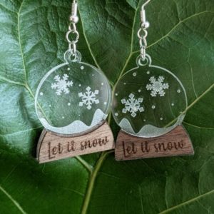 Snow globe earrings featuring etched snowflakes and wood base, laid on a green leaf.