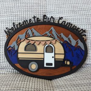 Rustic layered wooden sign with a scenic mountain backdrop and retro camper, reading 'Welcome to Our Campsite', displayed on textured fabric.