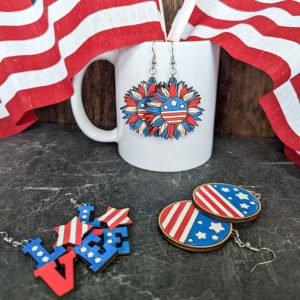 Fourth of July earring collection with American flag designs, displayed with red and white striped fabric and a white mug.