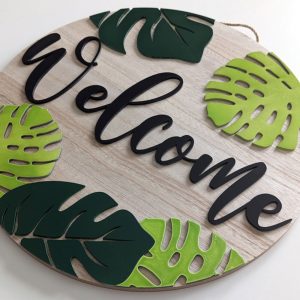 Angled view of a decorative wood sign featuring black 'Welcome' script and layered green monstera leaves in light and dark shades.