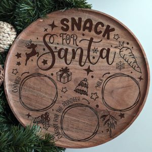 Engraved wooden plate labeled 'Snack for Santa' with designated sections for cookies, milk, and a carrot for the reindeer, surrounded by festive decorations like stars, a reindeer, and holly.