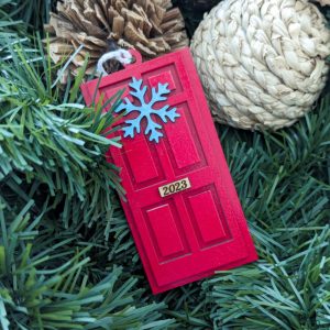 Single red door ornament labeled 2023 with a blue snowflake decoration, displayed nestled among pine branches, pinecones, and natural woven decor.