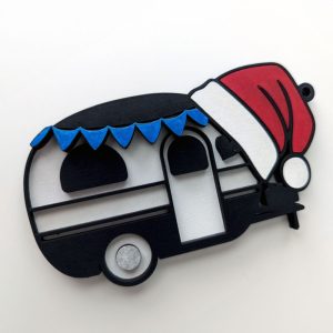 festive Christmas ornament shaped like a vintage camper trailer, decorated with a red Santa hat and a string of blue pennant flags. The ornament is placed on a white background.