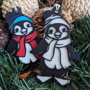Two laser-cut penguin Christmas ornaments, one with a blue hat and red scarf, the other with a white hat and scarf, placed on green grass with pinecones.