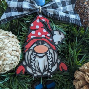 A single gnome ornament with a red polka dot hat, white braids, red mittens, and dangling blue boots, hanging on a green Christmas wreath. The gnome is topped with a black and white checkered bow and a blue wooden bead.