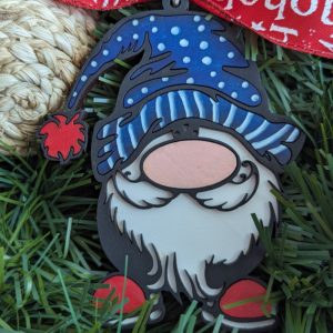 Christmas gnome ornament made of layered wood or acrylic, featuring a white beard, pink nose, and blue hat with white polka dots and stripes. The gnome wears red shoes and a red pom-pom on the hat. Displayed on green artificial grass with a red holiday ribbon in the background.