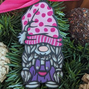 Close-up of a single gnome ornament with a pink polka dot hat and pink-striped brim. The gnome has a long gray beard and wears a purple dress with red and pink accents. It is placed on green pine garland with decorative natural accents around it.