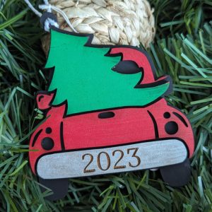 Laser cut red truck ornament with a green Christmas tree on top and "2023" engraving, placed on green holiday grass for display.