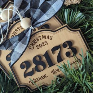 Rustic Christmas ornament with raised black ZIP code 28173 and Waxhaw, North Carolina text, decorated with a buffalo plaid bow and nestled in faux pine branches.