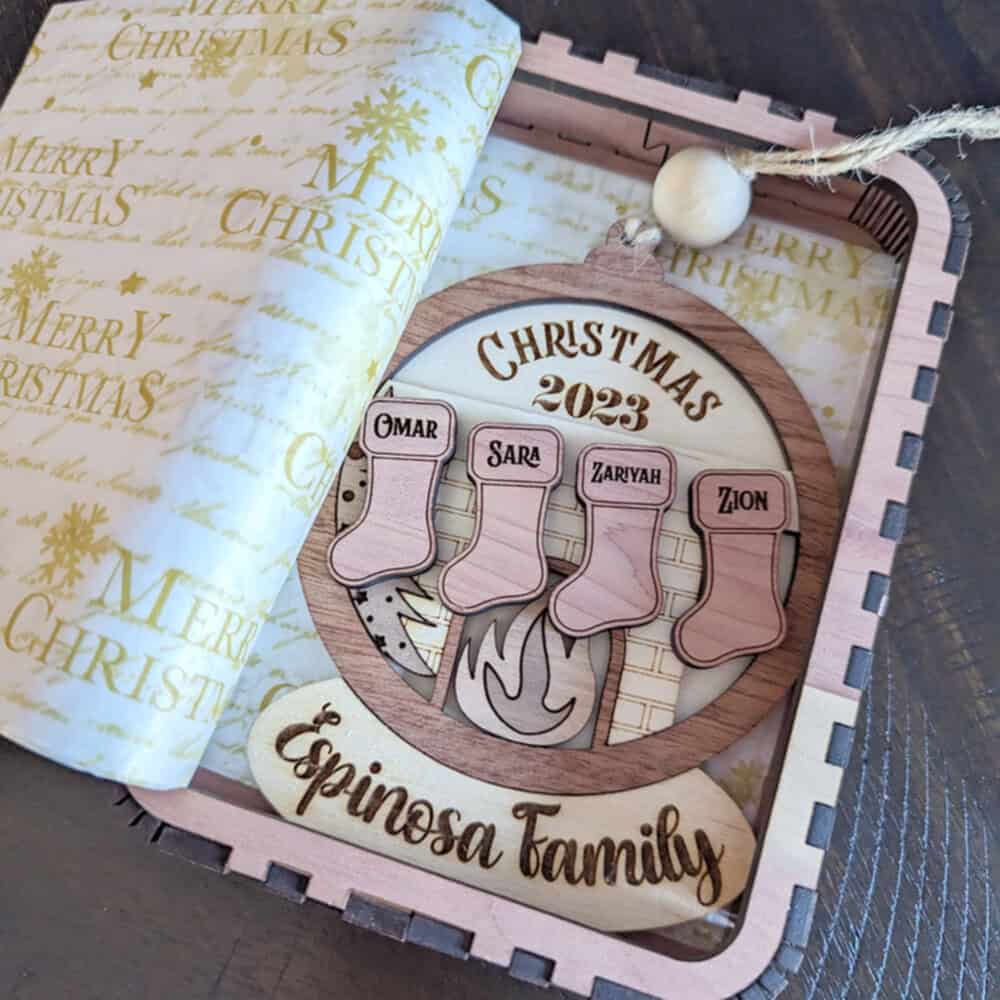 Personalized wooden Christmas ornament featuring four stockings labeled Omar, Sara, Zaryah, and Zion, hanging over a fireplace with 'Christmas 2023' engraved above and 'Espinosa Family' engraved below. The ornament is displayed in a decorative wooden gift box lined with festive 'Merry Christmas' wrapping paper.