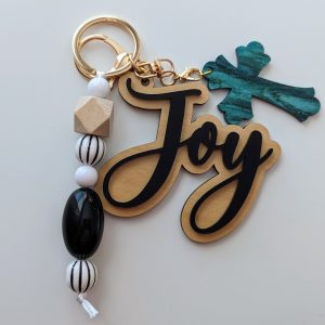 Keychain with "joy" script in black and gold, teal glitter cross charm, and black, white, and wood-tone beads with striped patterns, on a white background.