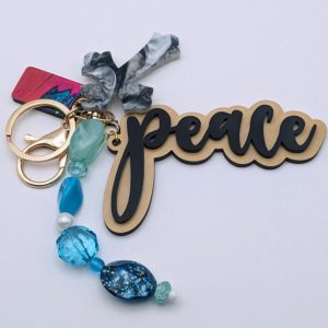 Keychain with "peace" text in black and gold, marbled gray cross charm, and blue-toned beads including a galaxy-patterned oval bead and translucent gems.
