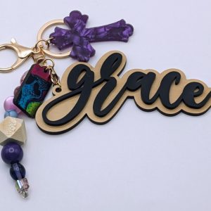 Keychain with "grace" text in gold and black, marbled gray cross charm, and turquoise and blue beaded strand including a galaxy-style oval bead.