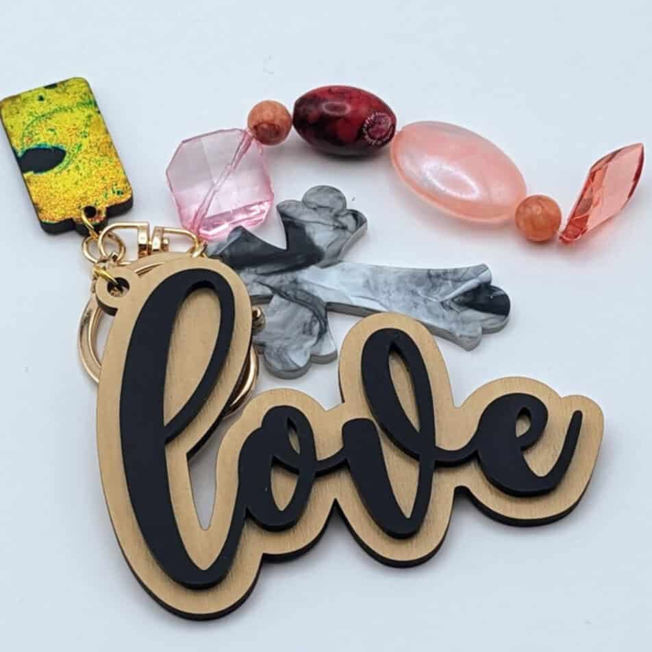 Keychain featuring "love" charm, gray marbled cross, and pastel-themed beads in pink, peach, and orange with a gold keyring.