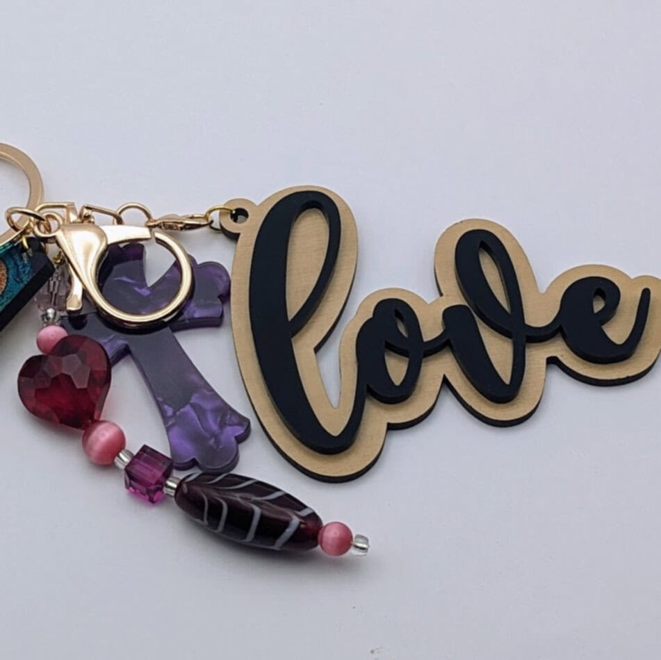 Keychain with gold and black "love" text, purple cross charm, and a strand of pink and purple beads including a red heart-shaped bead.