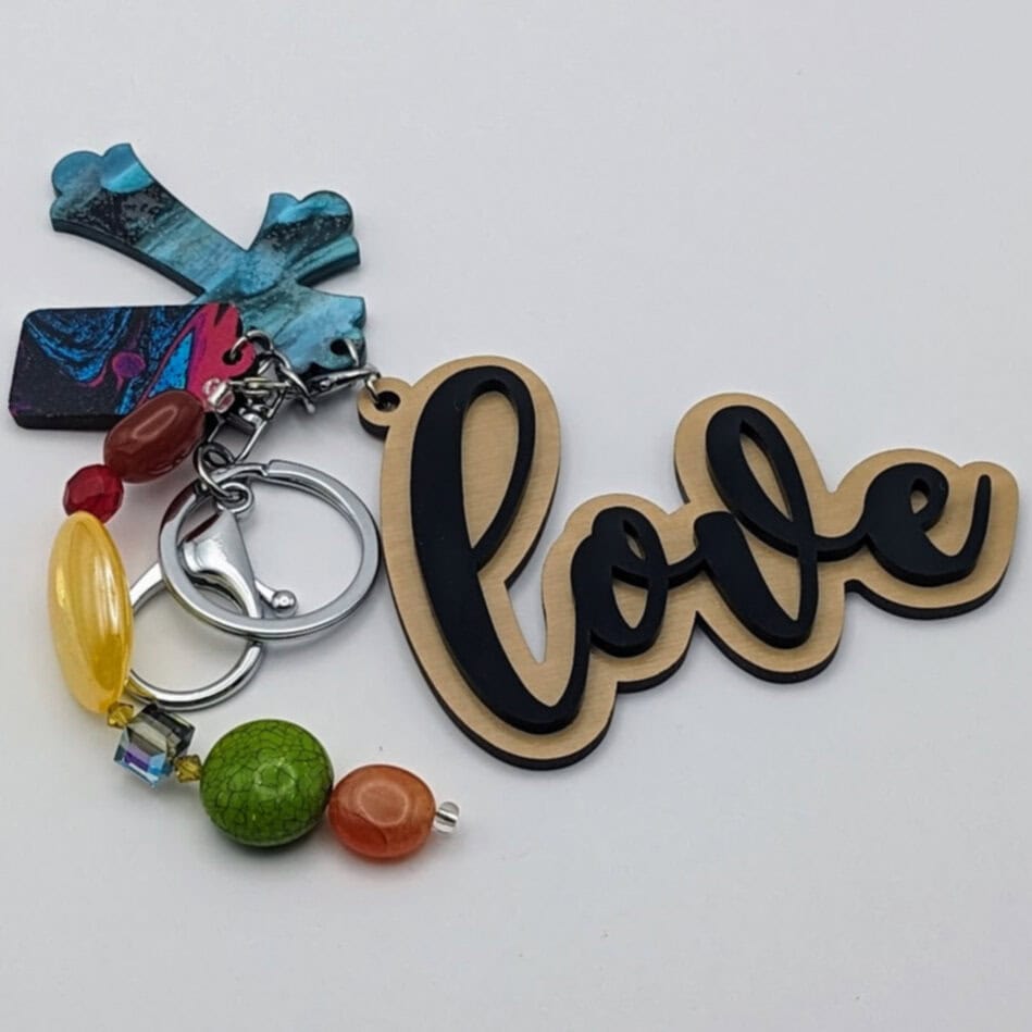 Keychain with "love" text in black and gold, blue marbled cross charm, and assorted bright beads in yellow, green, red, and orange.