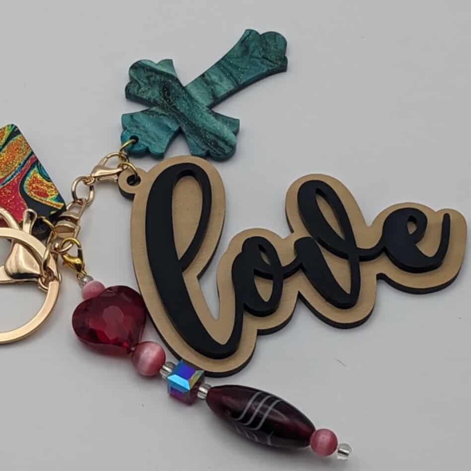 Keychain featuring "love" text in black and gold, teal green cross charm, and beaded strand with red heart and purple elongated bead.