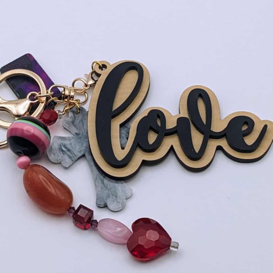 Keychain with "love" text in black and gold, attached to marbled gray cross charm and colorful beads including a red heart and striped round bead.