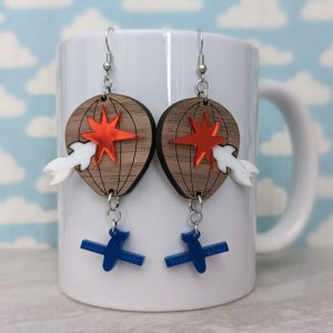 Hot air balloon-inspired earrings featuring layered wood with red starbursts, white rocket details, and blue satellite dangles, displayed on a white mug with cloud background.