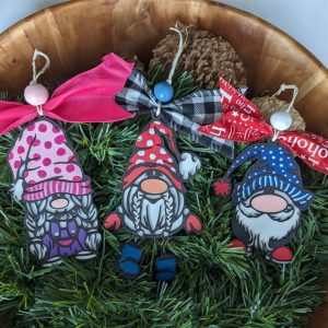Three gnome ornaments with red, pink, and blue hats displayed on green holiday foliage in a wooden bowl.
